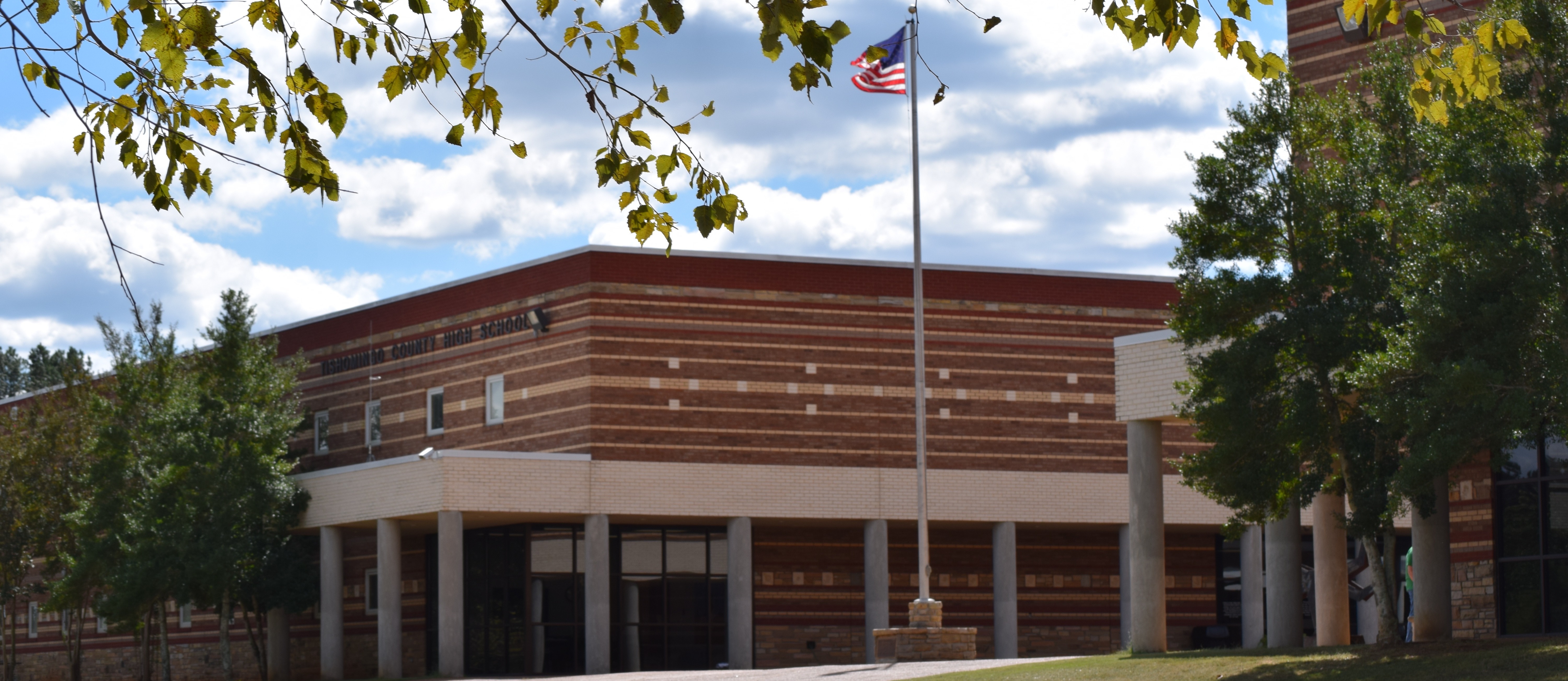The exterior front facade of Tishomingo County High School under a cloudy blue sky, featuring a modern patterned brick building with an American flag flying on a tall flagpole and tree branches framing the top of the view.
