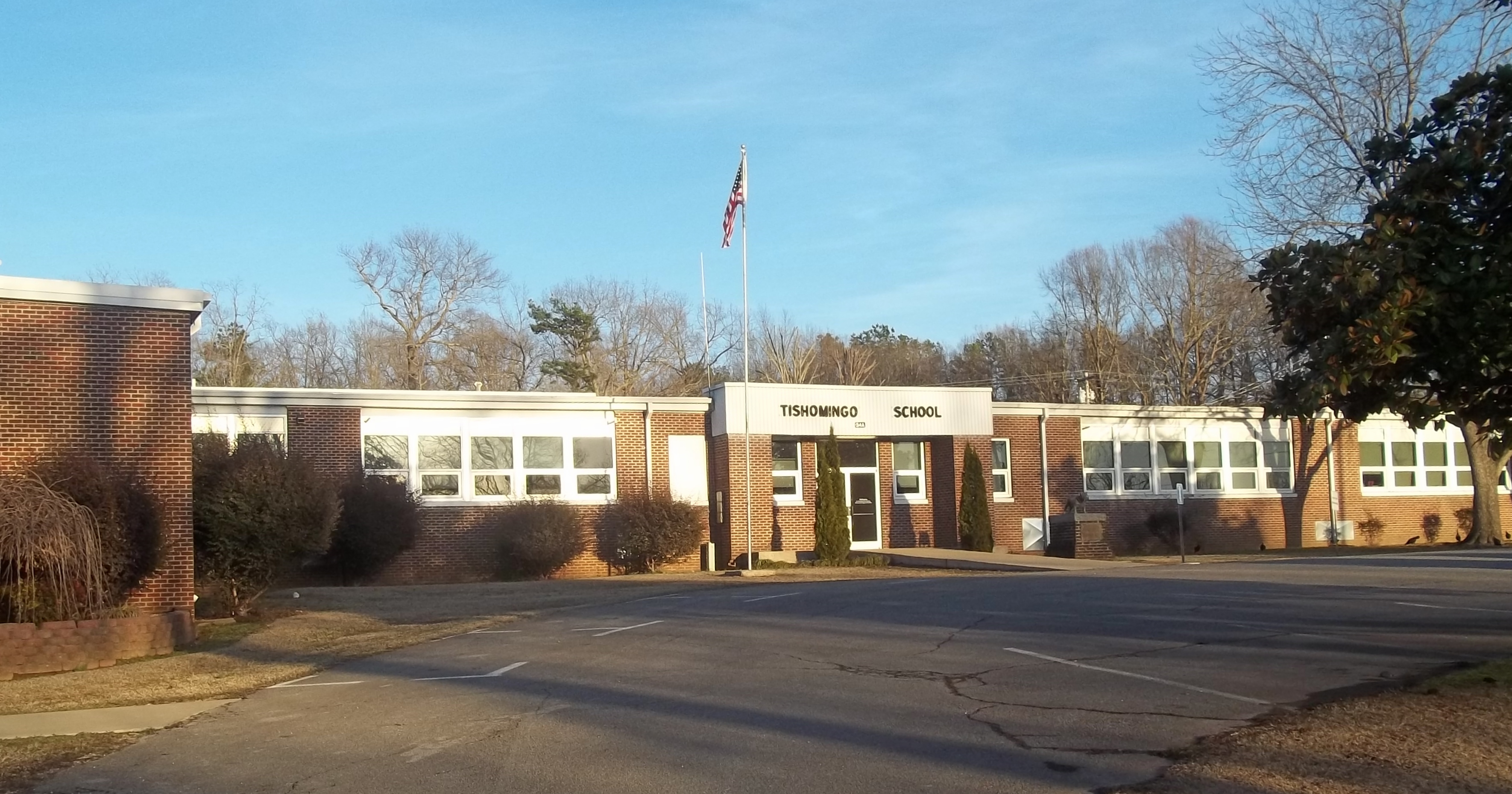 The front exterior of Tishomingo School, a single-story red brick school building featuring large banks of white-trimmed windows, a central glass entrance, and an American flag flying on a tall flagpole.
