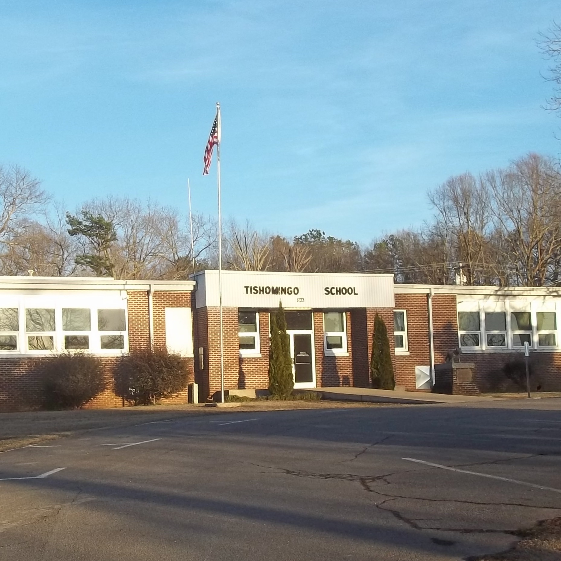 The front exterior of Tishomingo School, a single-story red brick school building featuring large banks of white-trimmed windows, a central glass entrance, and an American flag flying on a tall flagpole.
