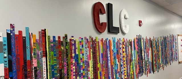 A long hallway wall displays a collaborative student art installation made of hundreds of colorful, vertically mounted painted wooden slats arranged side by side. Large three-dimensional letters spelling "CLC" are mounted above the artwork, and the corridor has white walls and a drop ceiling.