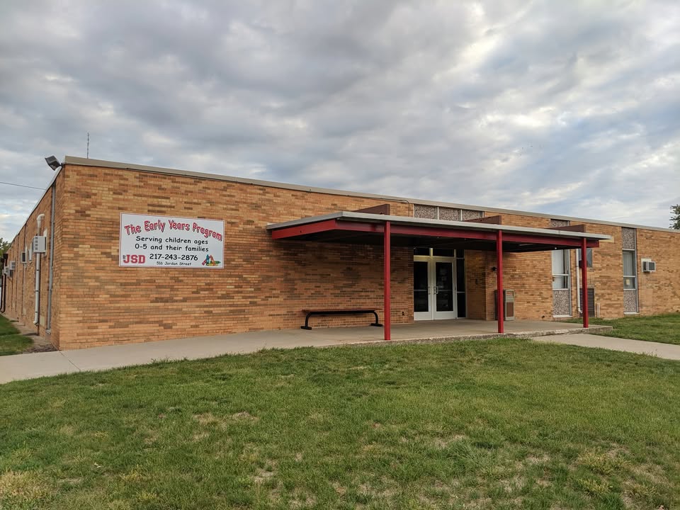 The Early Years Program building features a single-story brick exterior with a covered entrance supported by red columns. A sign on the front identifies the program as serving children ages 0–5 and their families, with a grassy lawn and cloudy sky in the foreground.