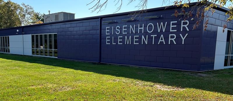 The exterior of Eisenhower Elementary School shows a modern building with dark purple and white panels, large windows, and the words "Eisenhower Elementary" mounted prominently on the side, surrounded by a grassy lawn and trees.
