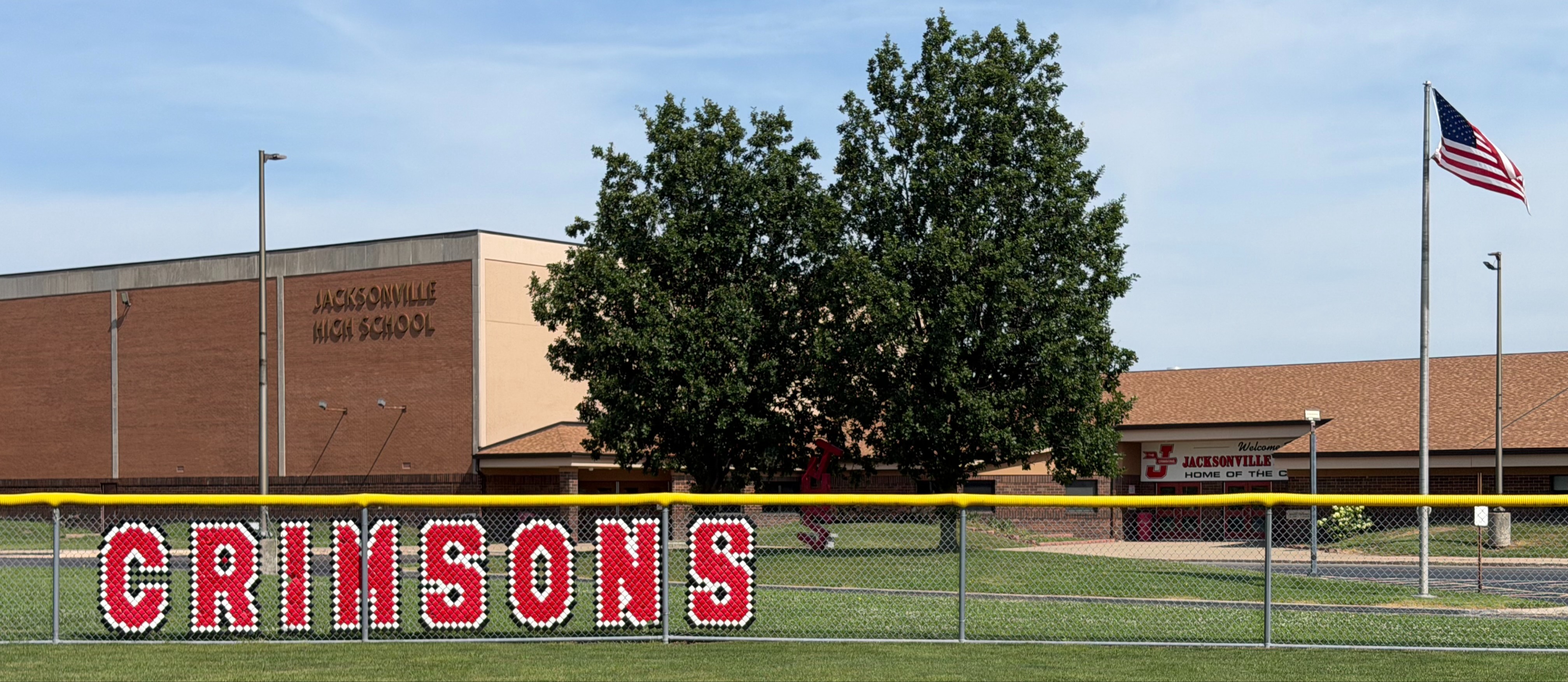 View of Jacksonville High School from behind a baseball or softball outfield fence displaying large red-and-white letters spelling "CRIMSONS." The brick school building, partially shaded by mature trees, sits in the background with the American flag flying on a flagpole near the main entrance. A grassy field and paved driveway are visible under a blue sky.View of Jacksonville High School from behind a softball outfield fence displaying large red-and-white letters spelling "CRIMSONS." The brick school building, partially shaded by trees, sits in the background with the American flag flying on a flagpole near the main entrance. A grassy field and paved driveway are visible under a blue sky.