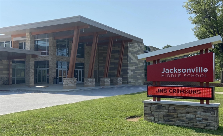 The entrance to Jacksonville Middle School features a modern building with large glass windows, stone columns, and angled wooden support beams. In the foreground, a red monument sign reads "Jacksonville Middle School," with an electronic display below showing "JMS Crimsons," surrounded by a green lawn on a sunny day.