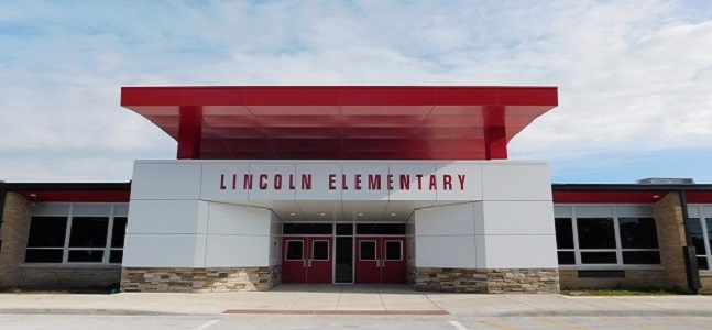 The main entrance of Lincoln Elementary features a modern white facade with a red canopy extending over the entryway. Red double doors sit beneath large letters spelling "Lincoln Elementary," with stone accents along the base and a partly cloudy sky overhead.