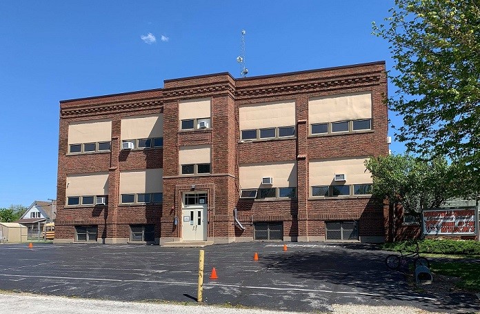 The exterior of Murrayville-Woodson Elementary School features a three-story red brick building with rows of windows, a central entrance, and a paved parking area in front. A school sign is visible near the entrance, with trees framing the building under a clear blue sky.