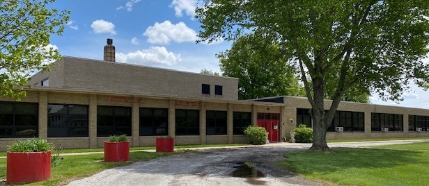 The exterior of North Elementary School features a long, single-story brick building with large windows, red entrance doors, and mature trees shading the front lawn. A sidewalk leads to the main entrance, with landscaped planters and a clear sky in the background.