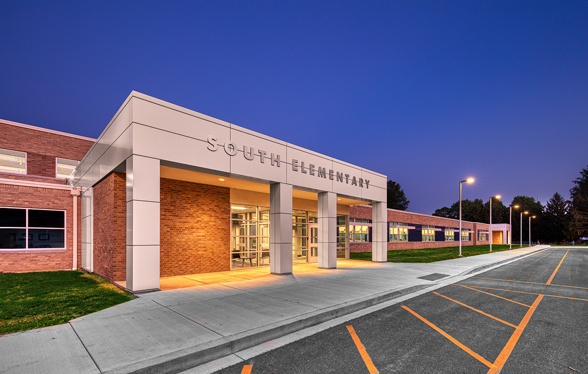 The main entrance of South Elementary features a modern brick building with a white canopy supported by square columns. The words "South Elementary" are displayed above the glass entrance, with illuminated walkways and a parking area at dusk.