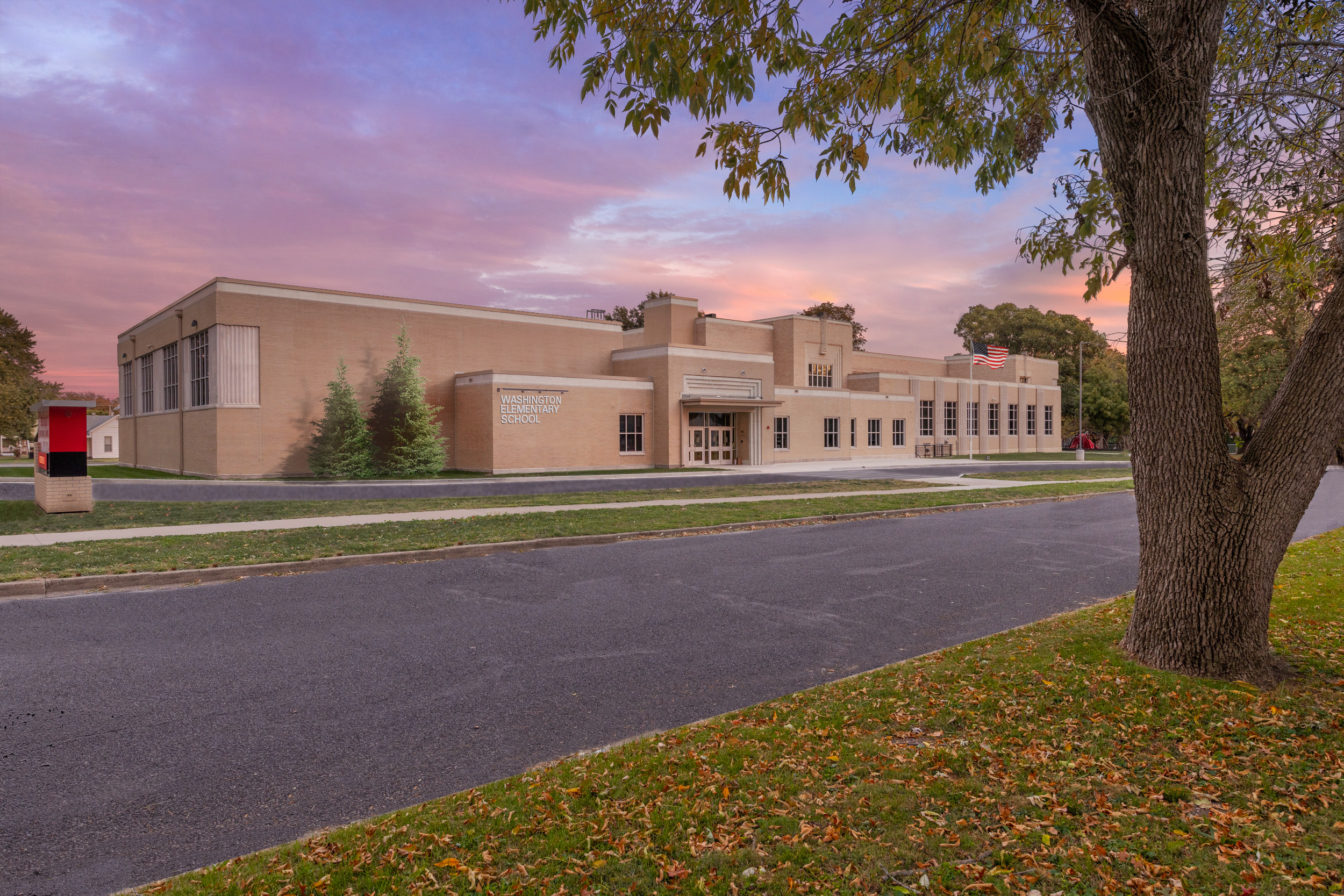 A wide view of Washington Elementary School shows a tan brick school building with an Art Deco-inspired entrance and large windows. An American flag flies near the front entrance, while a large tree frames the foreground beneath a colorful sunset sky.