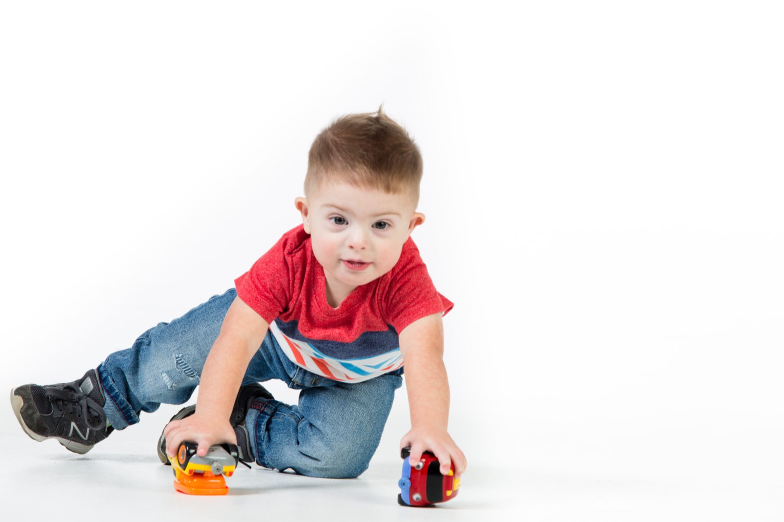 A young child in a red shirt and jeans playing with two colorful cars on a white floor.