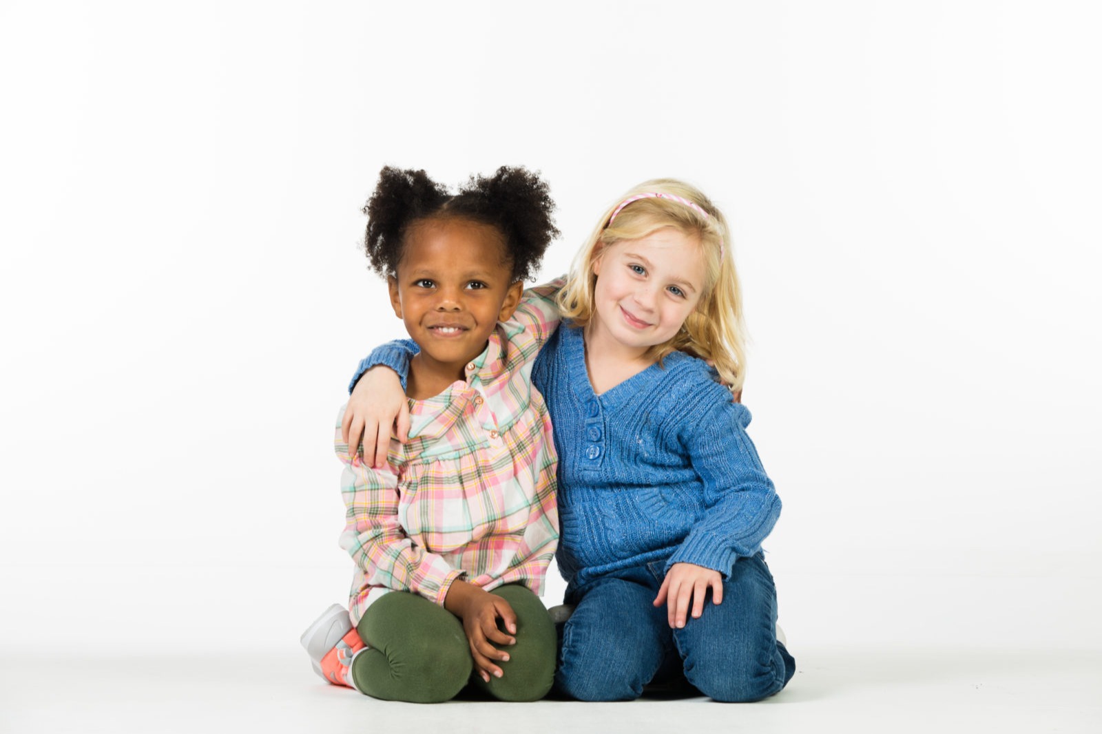 Two girls sitting on a white floor, one wearing a plaid shirt, the other in a blue sweater, smiling and posing.
