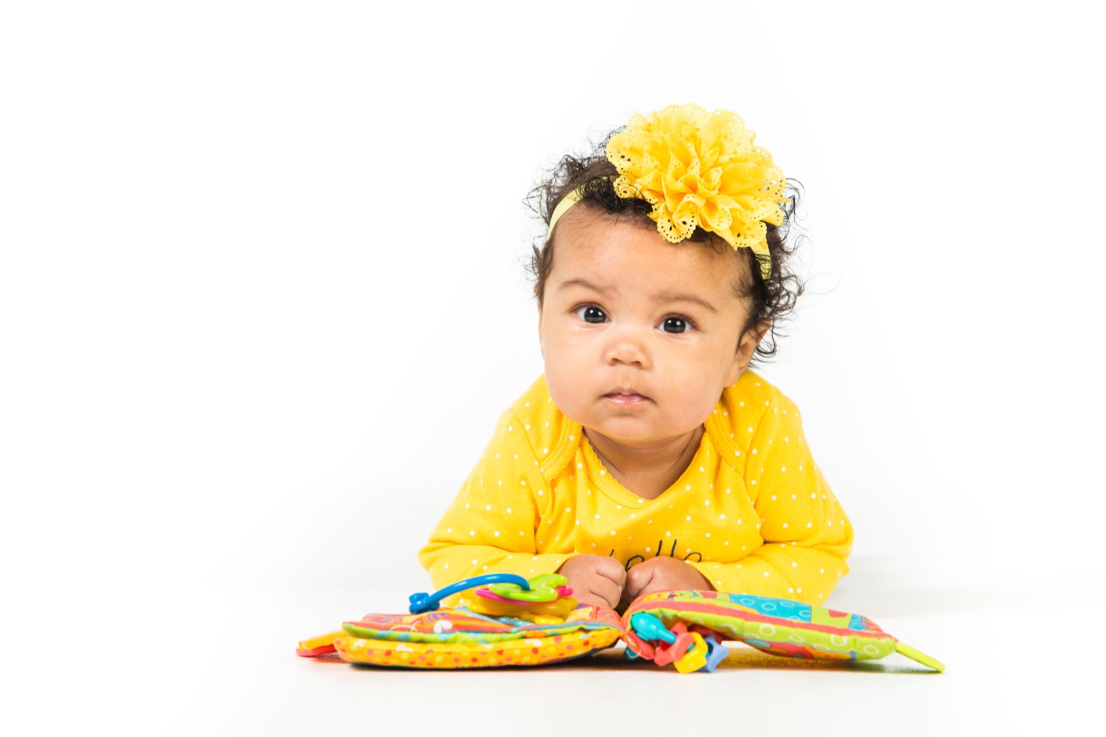 Infant girl with curly hair sits on floor with a yellow headband, a yellow shirt, and a colorful book.