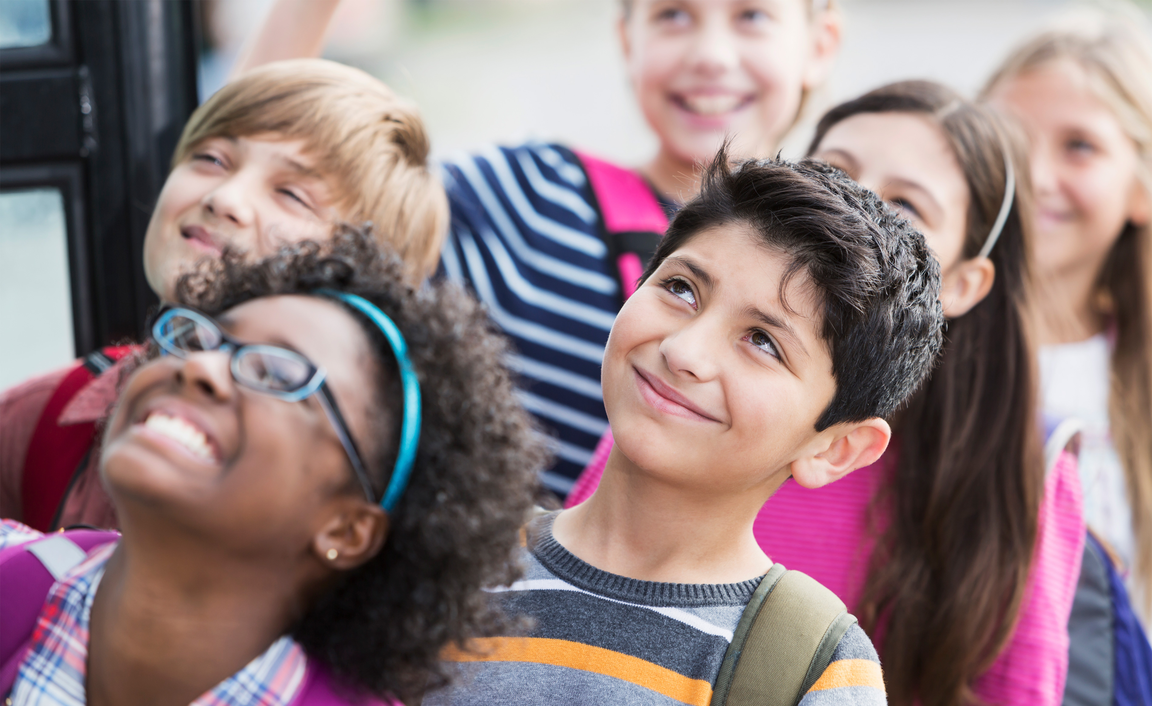 A group of children, some with glasses, wearing backpacks, standing together and smiling.