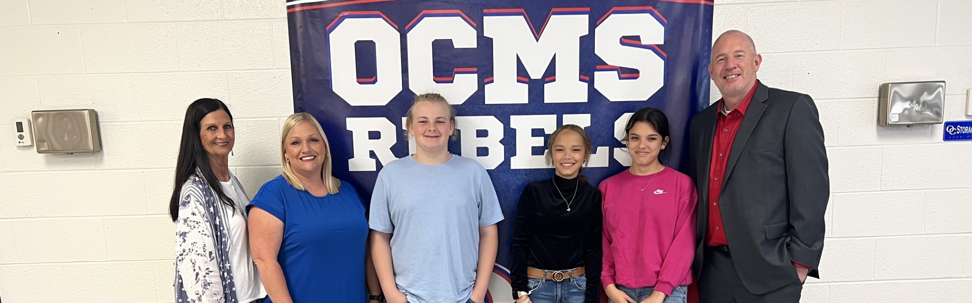 OCMS Admin and students smile in front of OCMS sign.