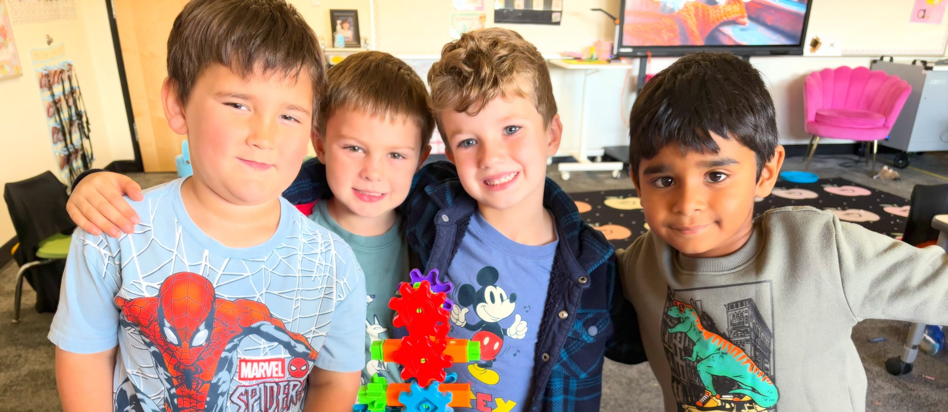 Four young boys stand together in a classroom playing with colorful interlocking gears they built with toy construction pieces.