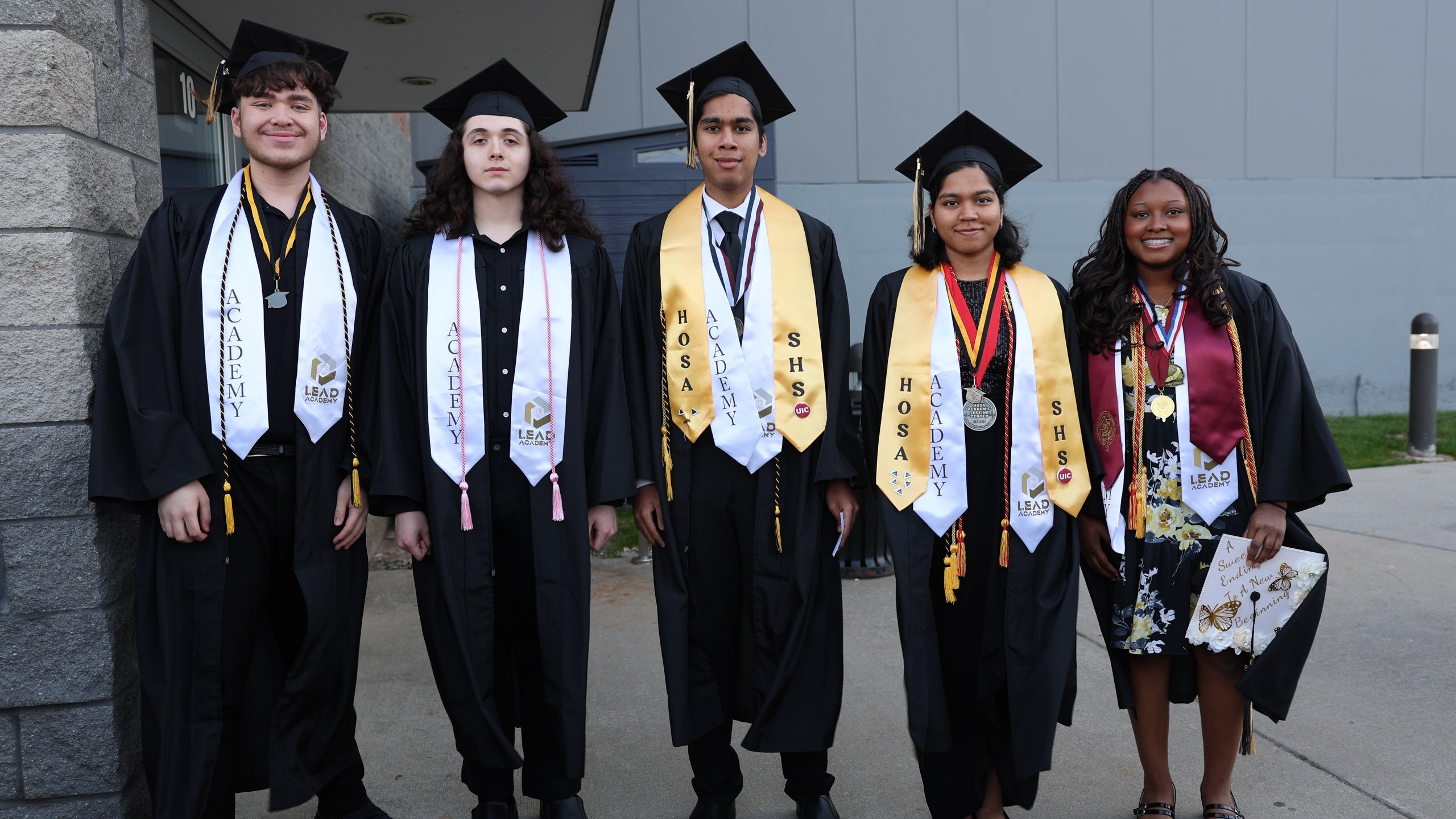 Five young adults in graduation robes pose for a photo outside a building. 