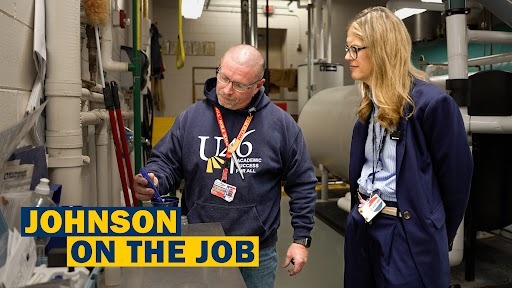 A man and woman in blue jackets discuss something in an industrial area with a large pipe.