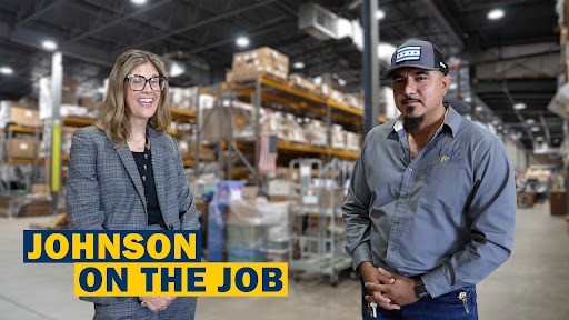 Two people, a woman and a man, are standing in a warehouse with shelves filled with boxes.