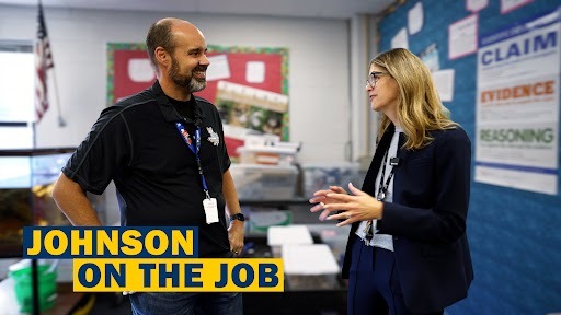 Two people, one with a nametag reading "Johnson", speak in an office. A blue bulletin board is in the background.