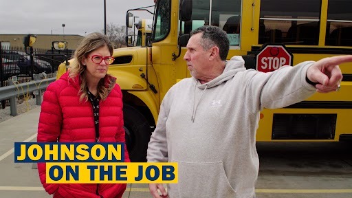A man and woman stand in a parking lot with a yellow school bus behind them.