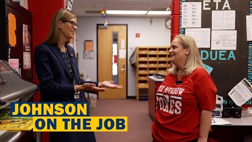 Two women stand in an office; one wears a blue blazer, and the other wears a red T-shirt.