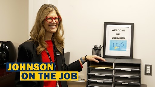 A woman wearing glasses and a red top stands behind a desk, pointing at a welcome sign.