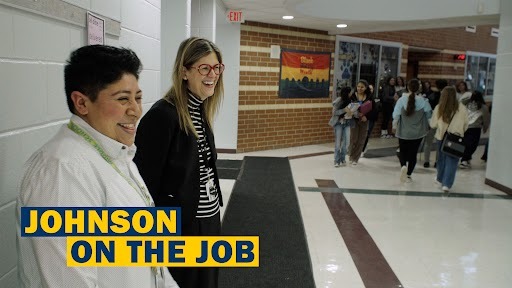 Two people stand near a wall with a sign that says Johnson on the Job. Behind them, students walk in a hallway.