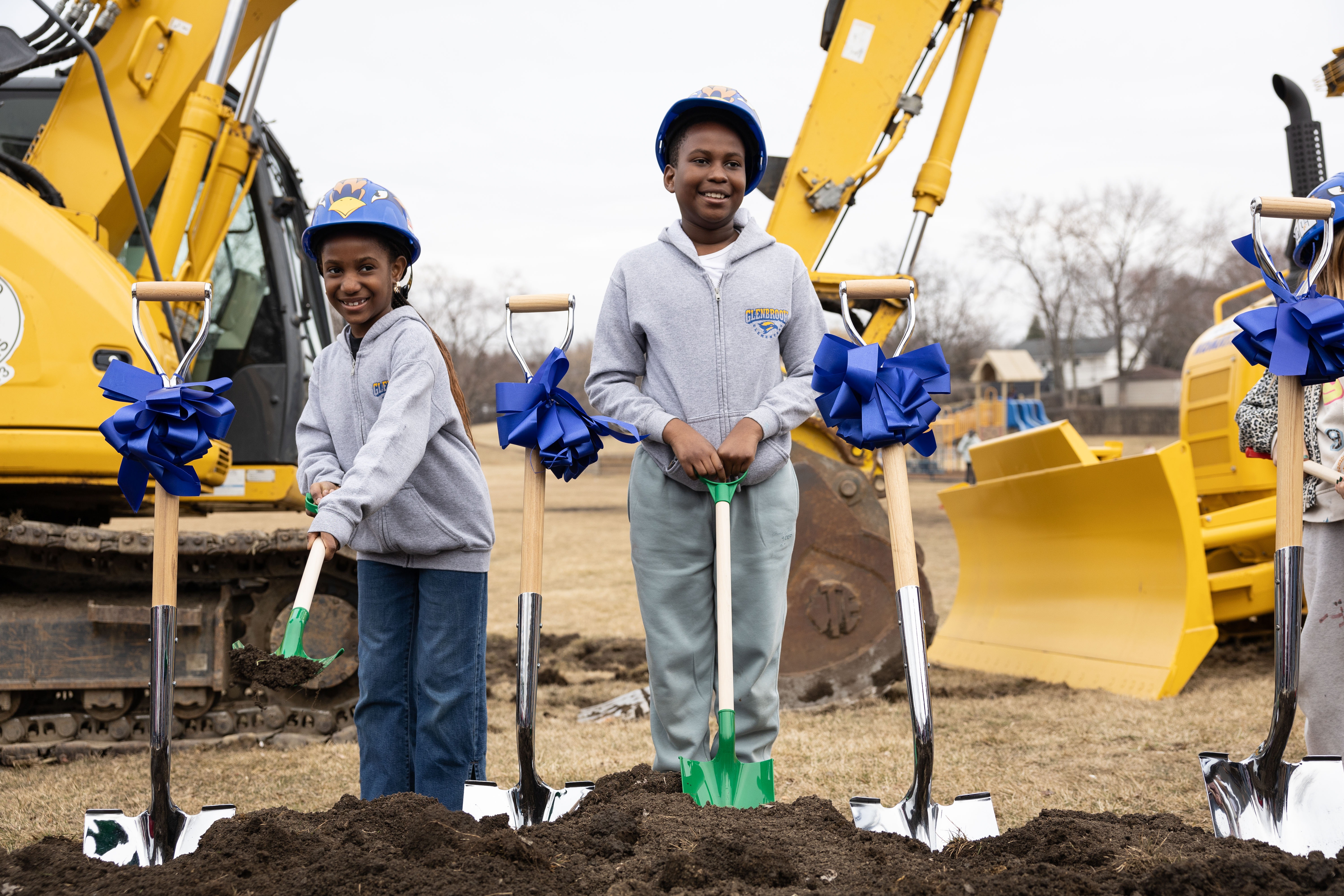 Three children stand in a hole holding shovels, with a yellow excavator and blue ribbons in the background.