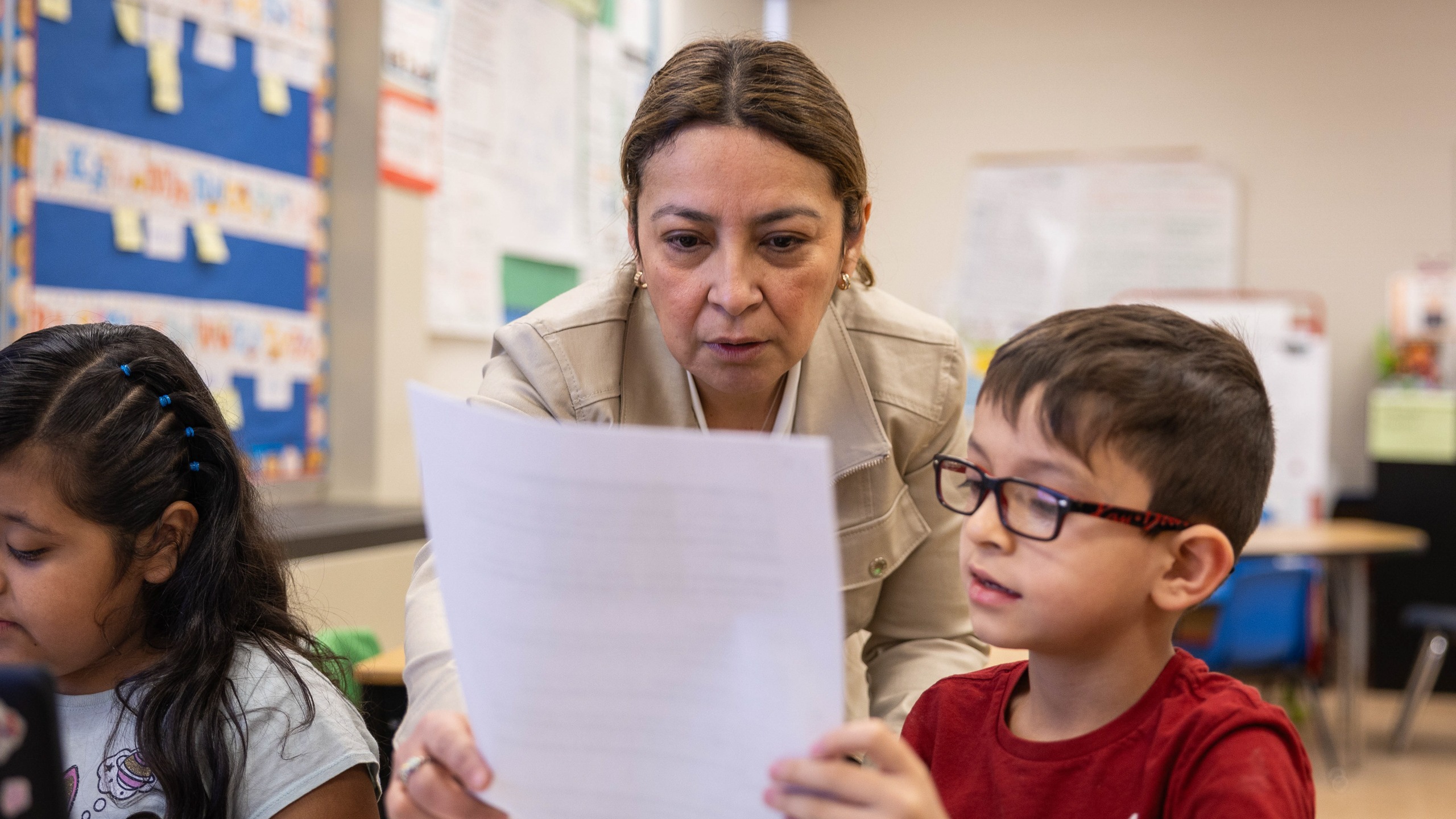 A teacher looks over a paper held by a young boy while a girl watches; classroom walls have decorations.