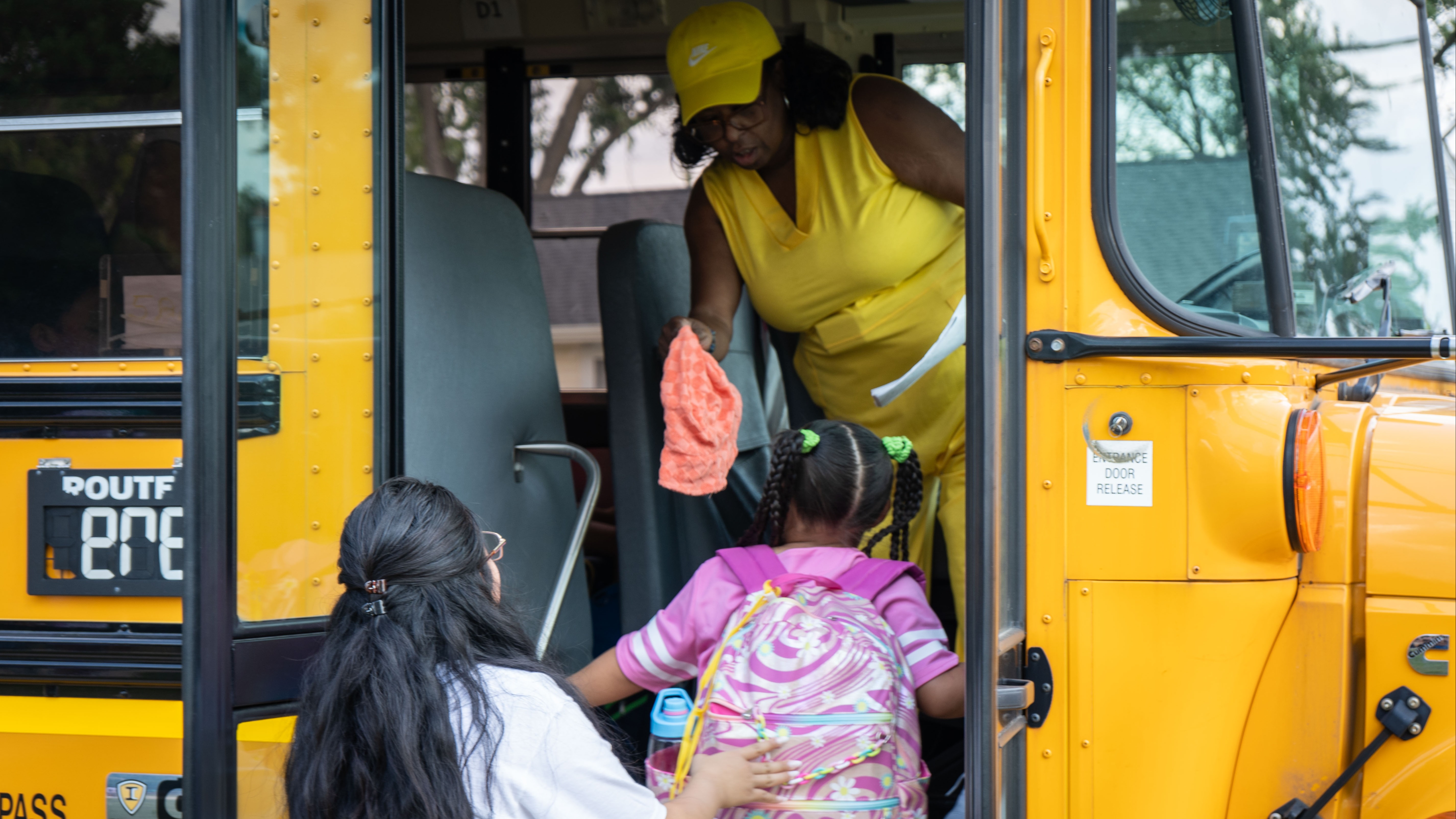 A woman in yellow helps a young girl with a backpack board a yellow school bus.