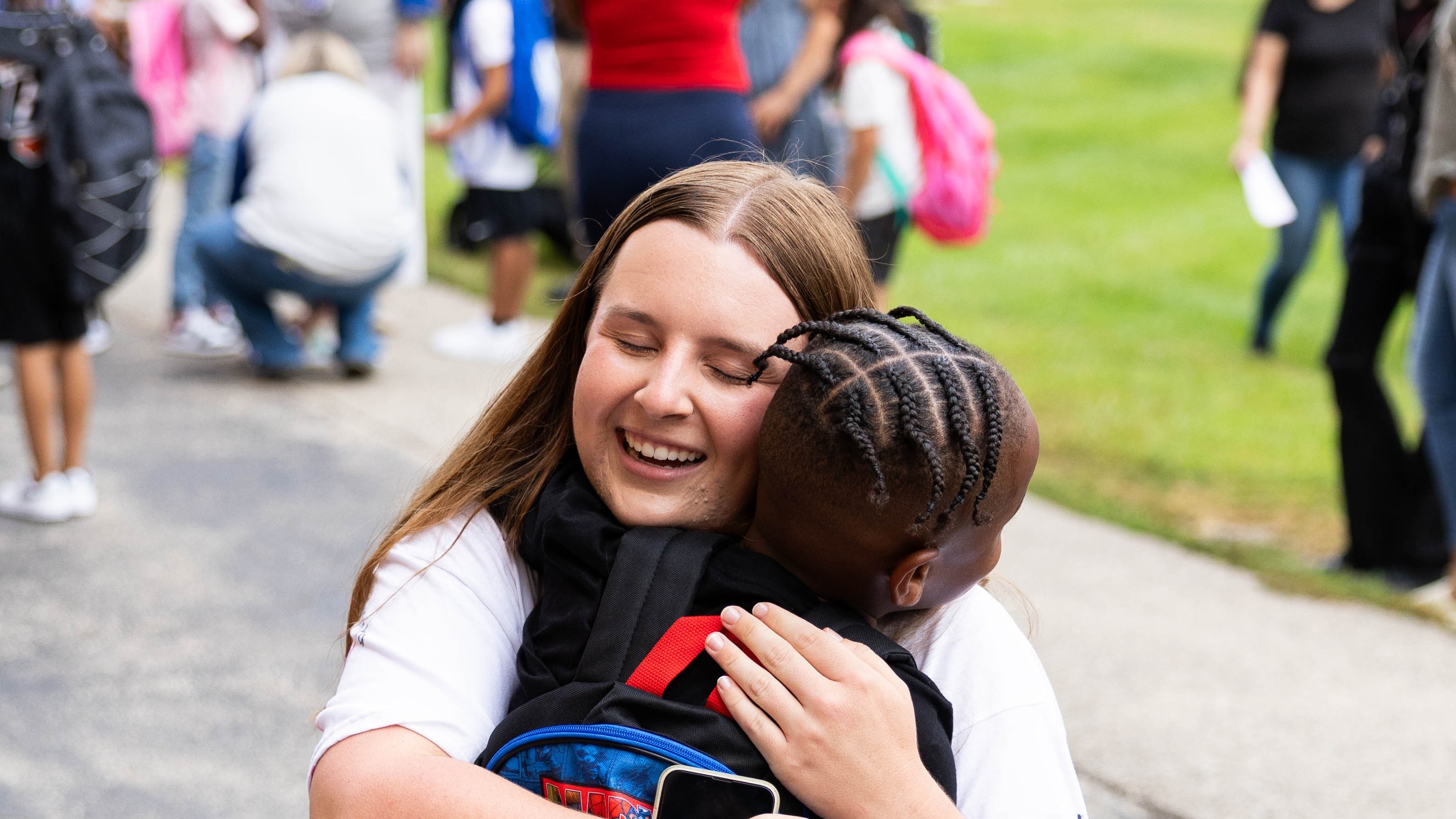 A young woman kneels on a road, embracing a child wearing a superhero backpack. They are surrounded by other people.