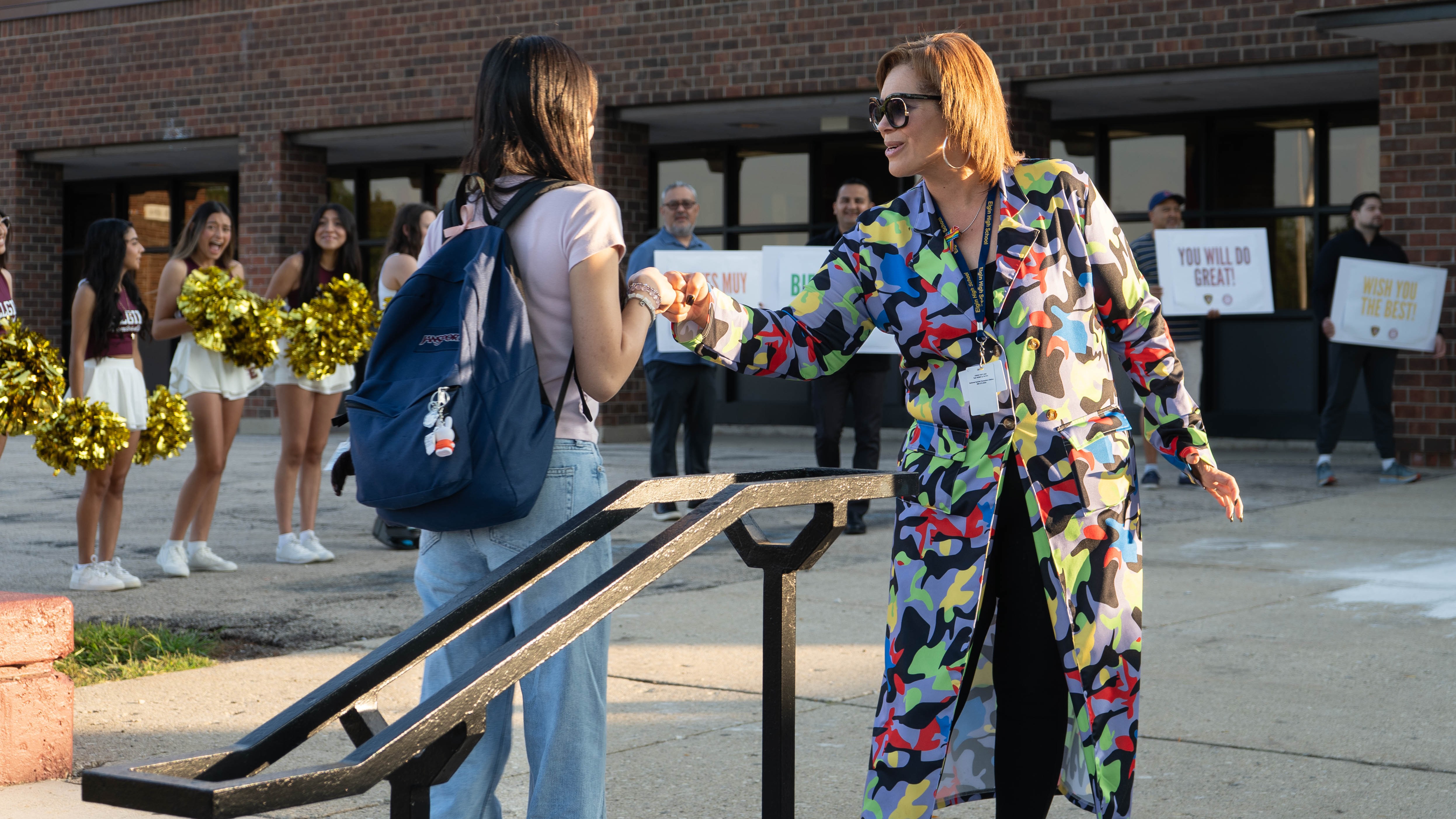 A woman in a colorful outfit shakes hands with another woman. Behind them, cheerleaders stand with pom-poms, and a brick building.