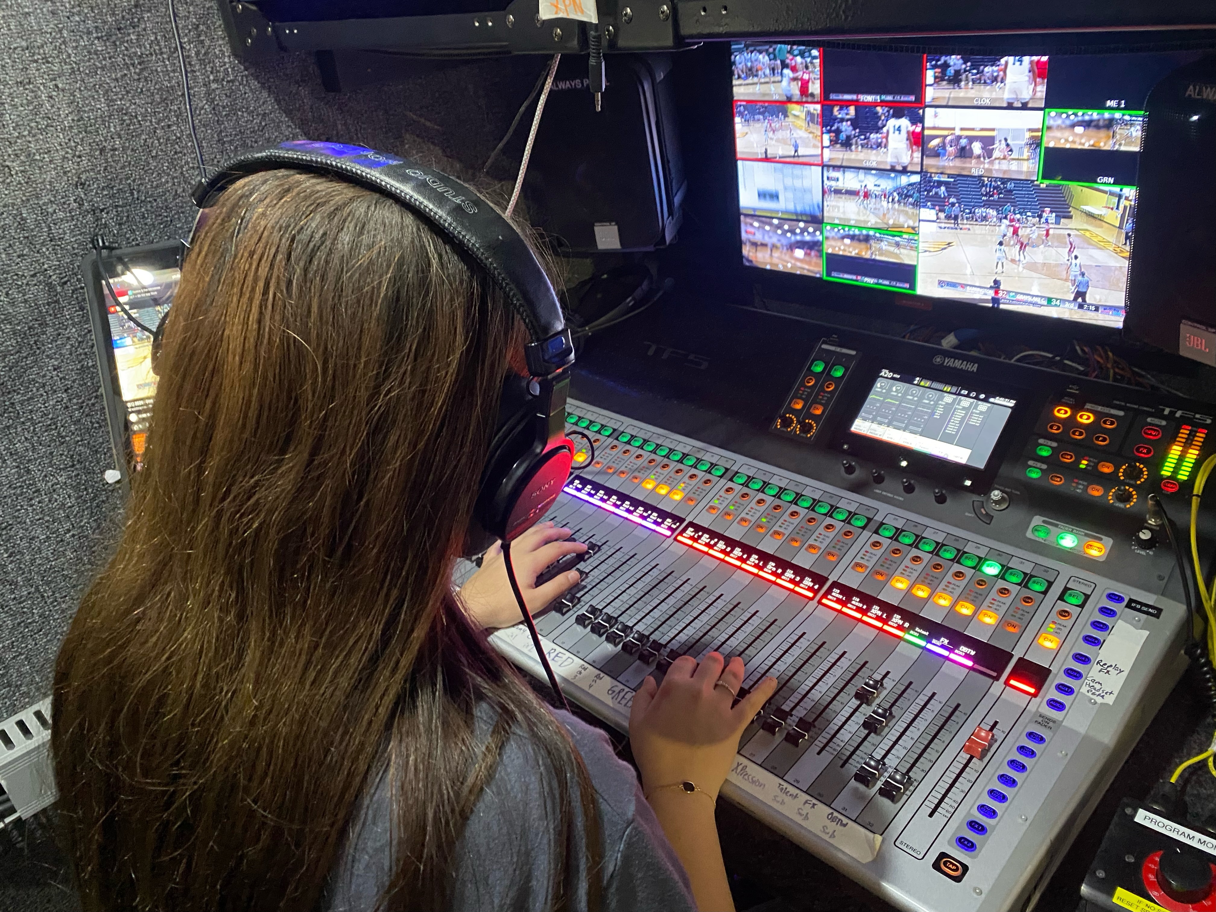 Person sitting at a mixing board, using a computer, wearing headphones. Monitors showing sports footage.