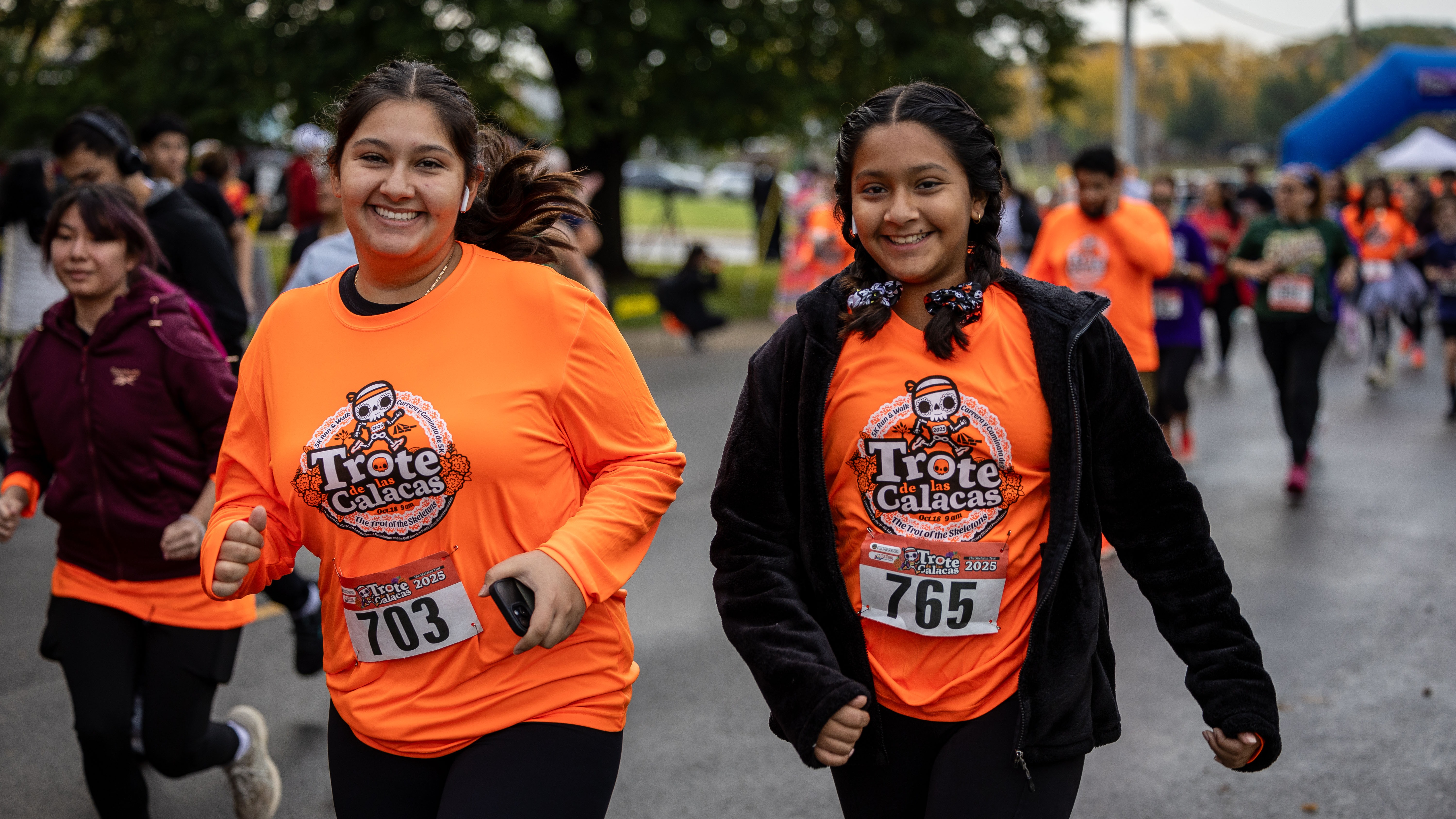 Participants in orange shirts running in a race. Background features blurred runners, trees, and an arch.