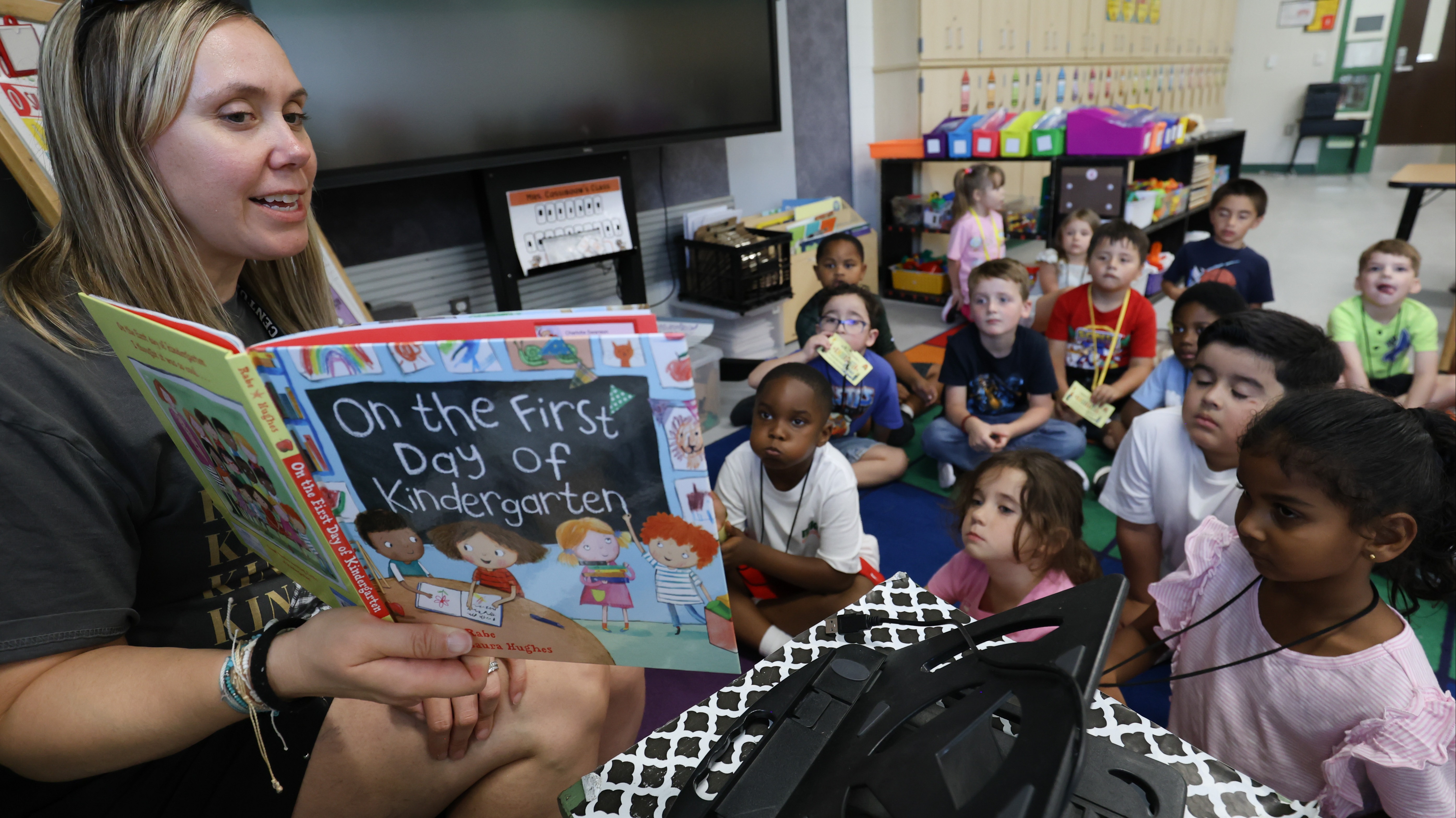 A woman reads a book to a group of children seated in a classroom. A screen and furniture are in the background.