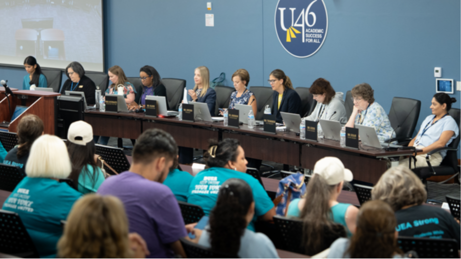 The School District U-46 Board of Education members sit at a long table with laptops for the Board meeting. The U-46 logo is behind them on the wall.