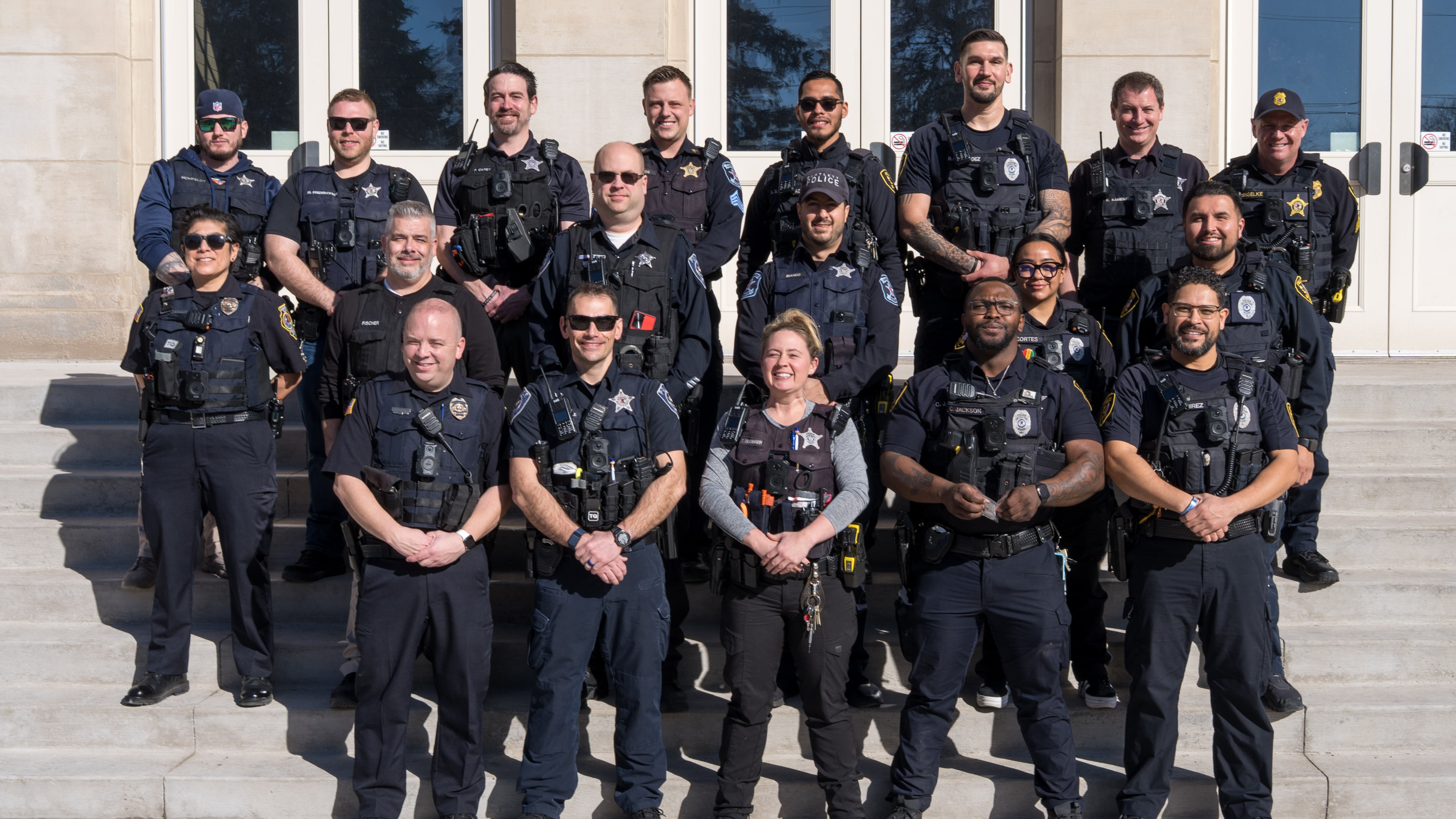 A group of police school resource officers standing on steps of the U-46 Educational Services Center. They are wearing police officer uniforms .