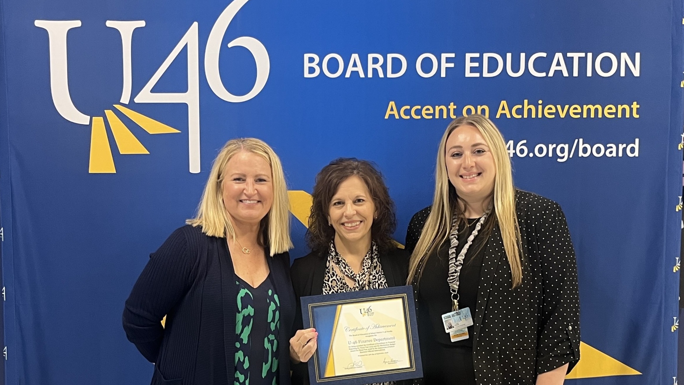 Three women stand next to each other in front of a blue backdrop with U46 Board of Education.