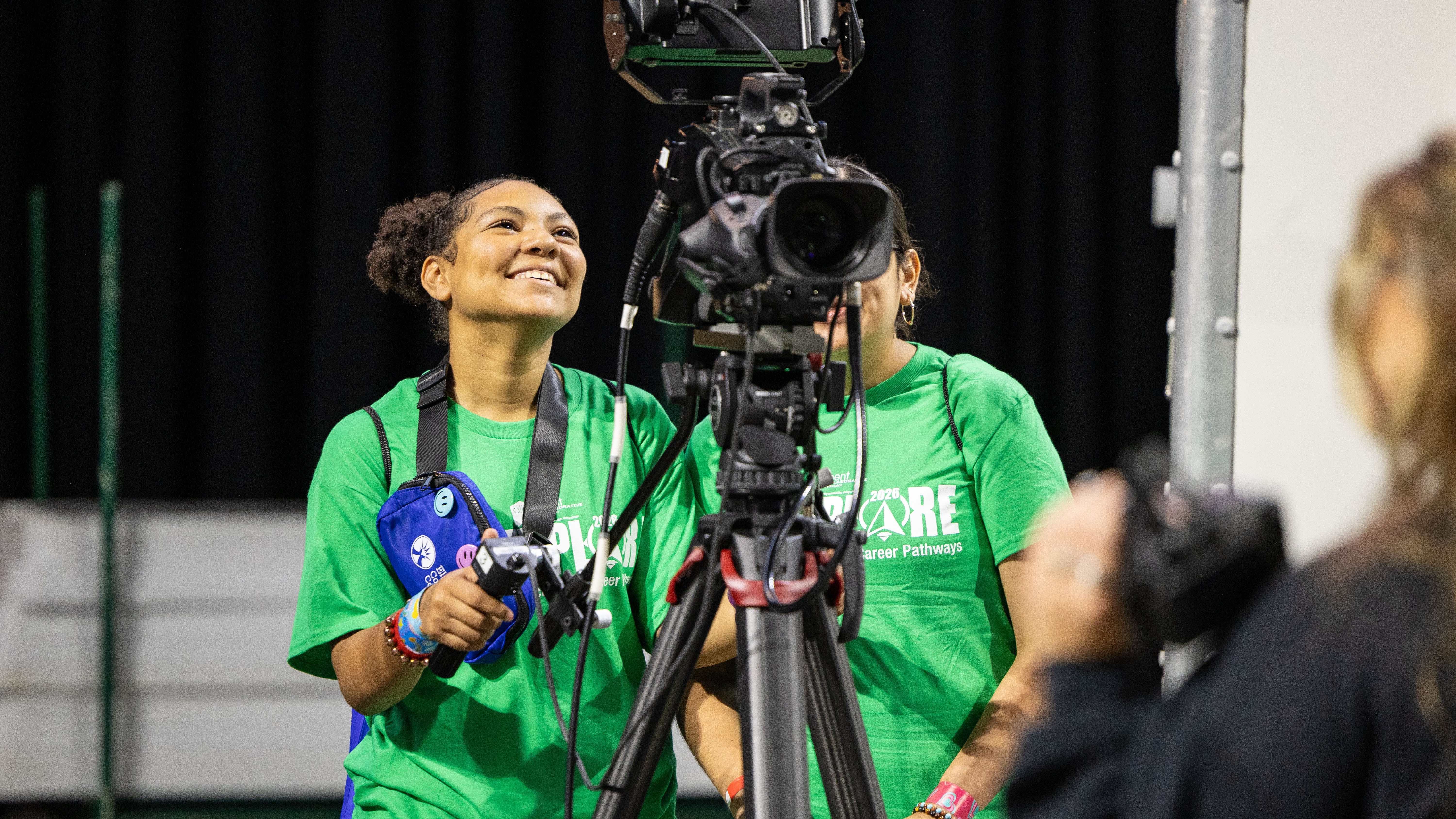 Three people in green shirts and black pants are near a tripod with a camera on it.