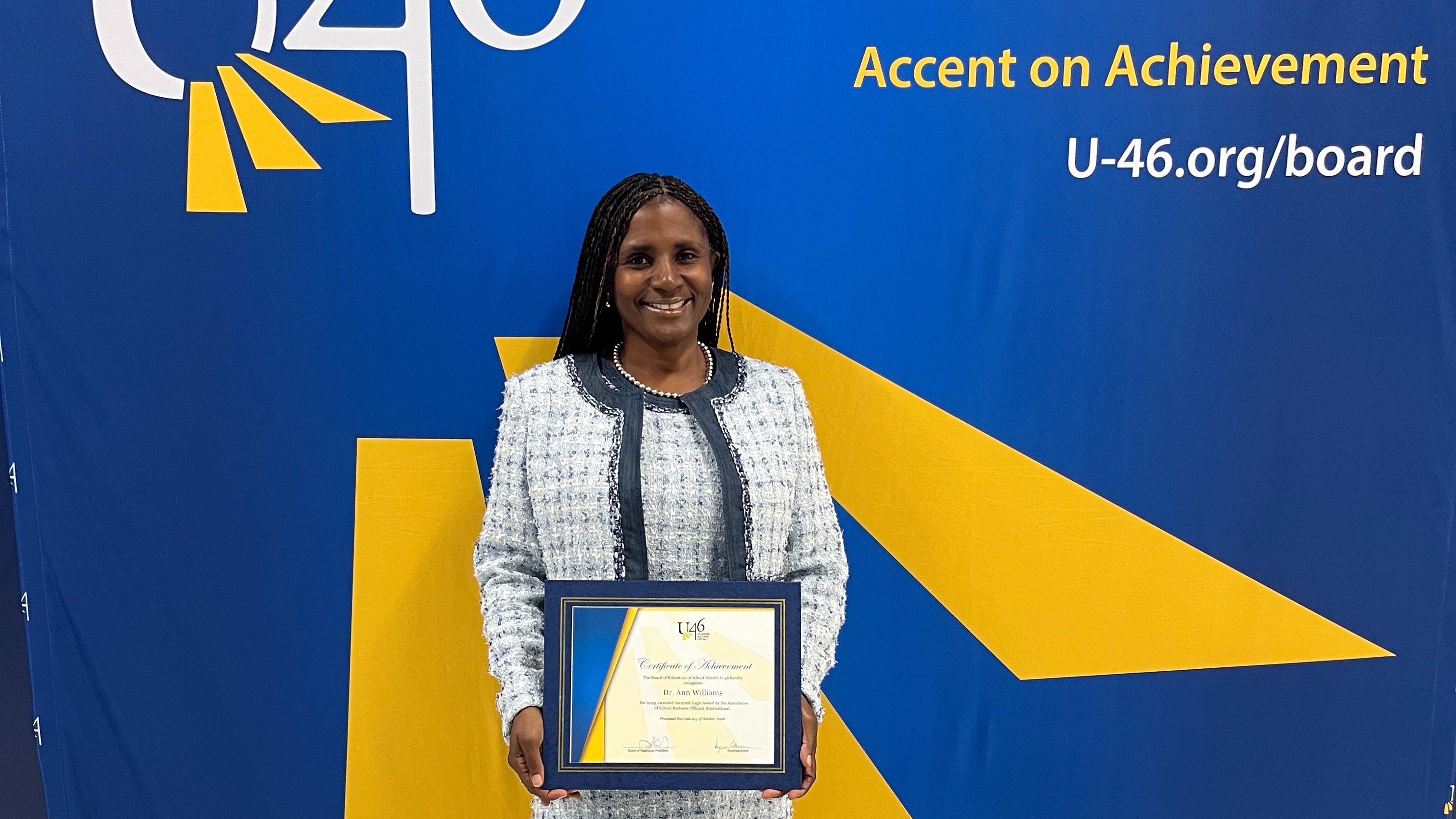 A woman stands in front of a blue backdrop with a yellow accent. She holds a framed award.
