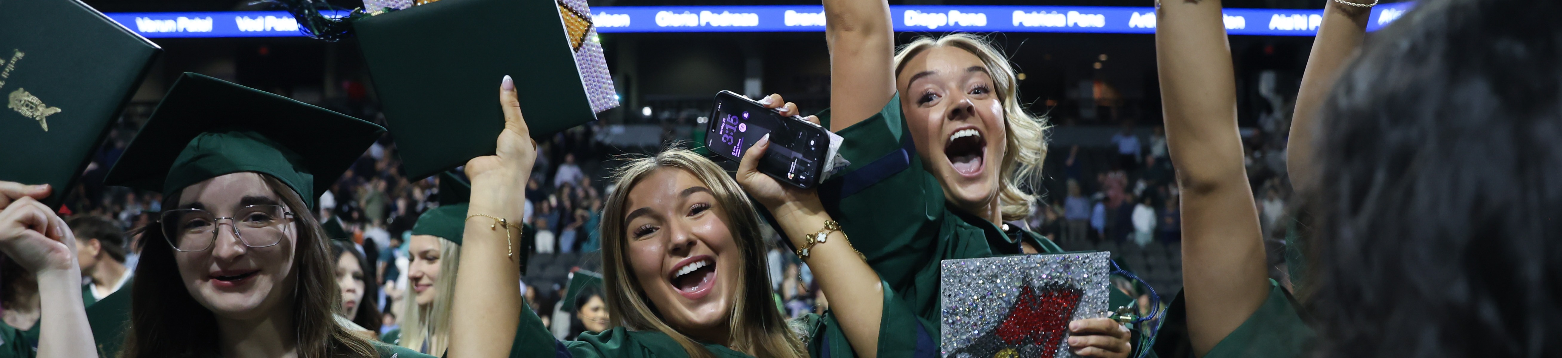 Graduates in green gowns celebrate at a ceremony. They hold caps and phones, and one holds a sign.