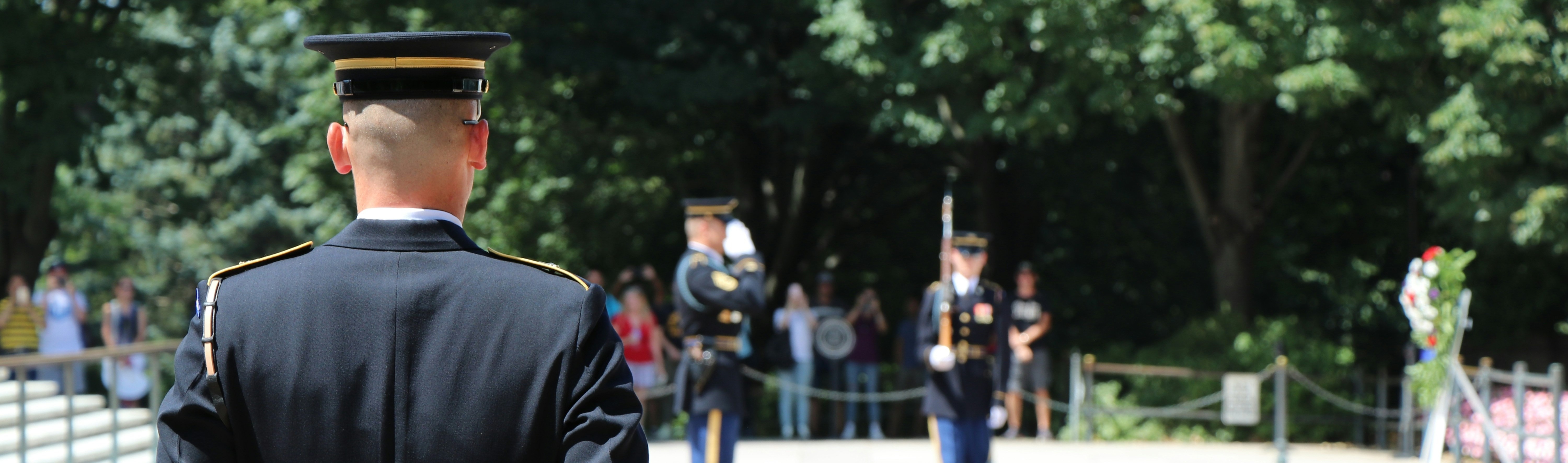 Soldiers in uniform stand on a concrete platform with trees in the background. One soldier faces away.
