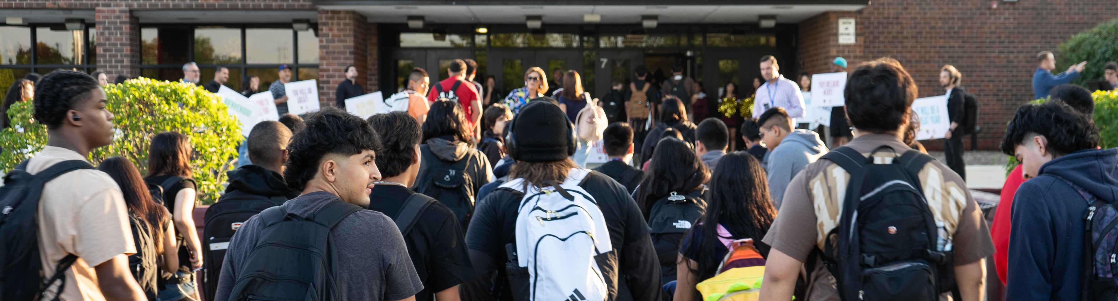 Students wearing backpacks walk toward the entrance of a brick building with trees and shrubs.