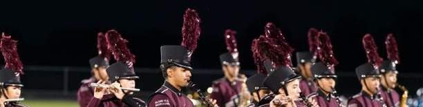 A marching band performs on a field at night. They wear maroon uniforms and black shoes, holding various instruments.