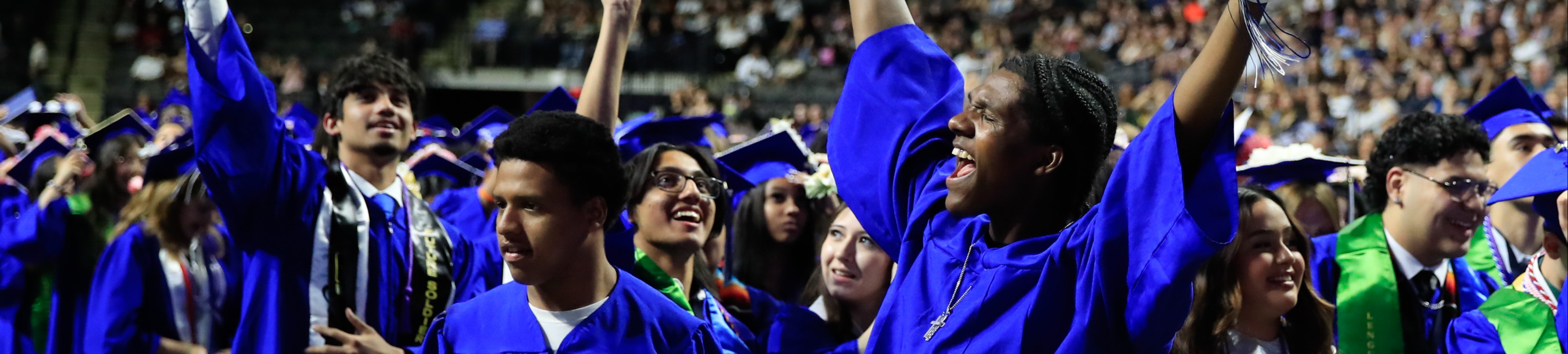 Graduates in blue robes stand, some holding diplomas, in a stadium; others cheer and watch.