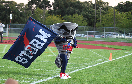 A mascot in a cone costume holds a blue flag with white text on a football field.