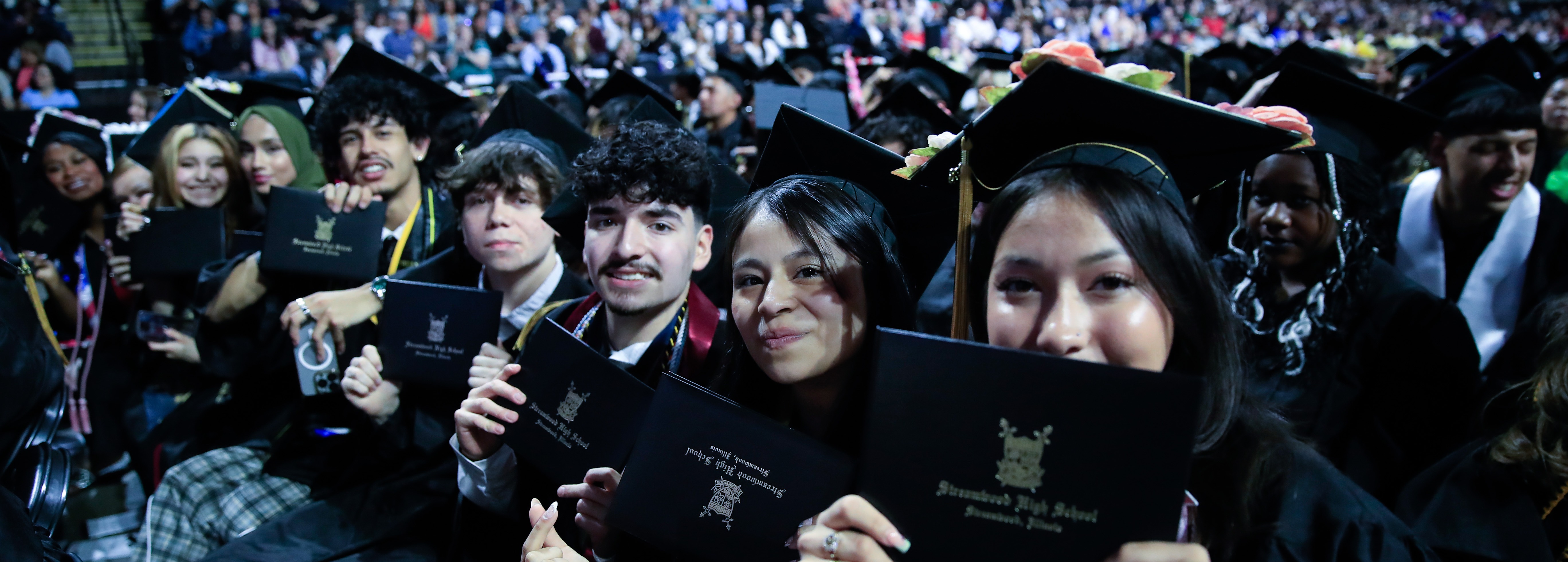 Graduates wearing caps and gowns sitting in a stadium, holding diplomas and smiling at the camera.