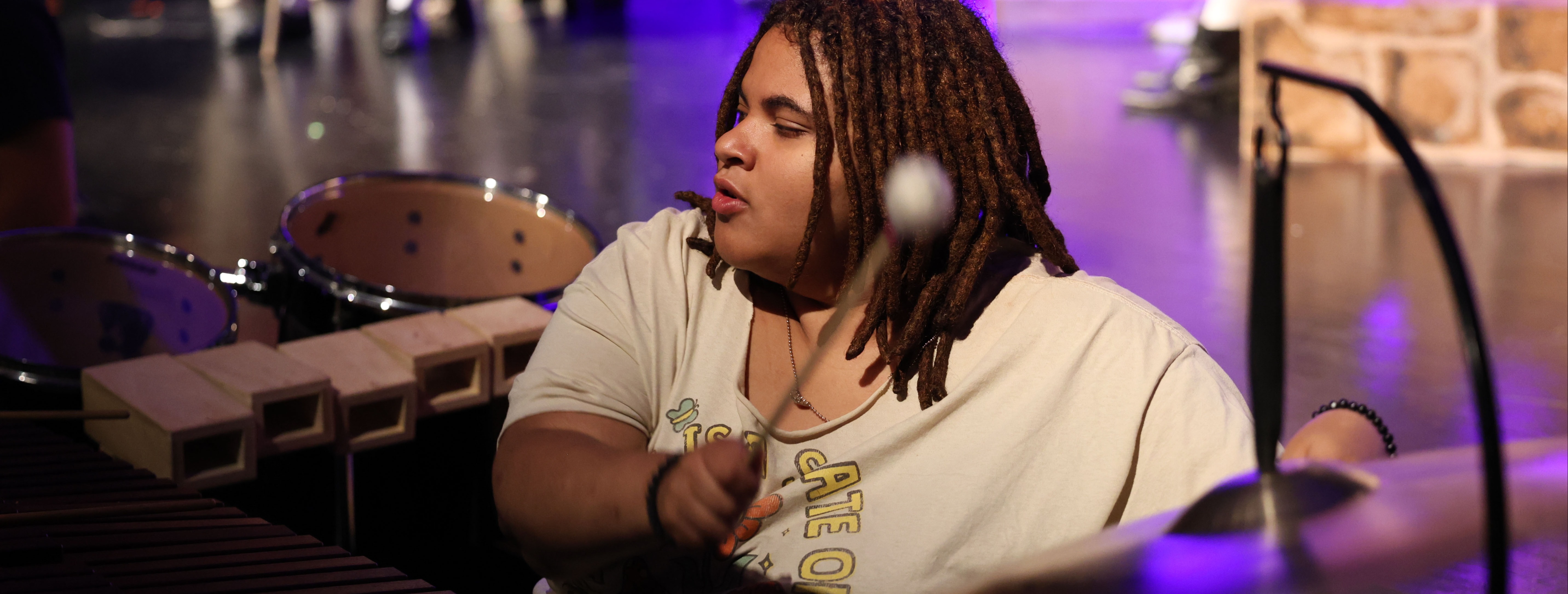 A person plays the xylophone and drums in a performance. They have curly hair and wear a white shirt.
