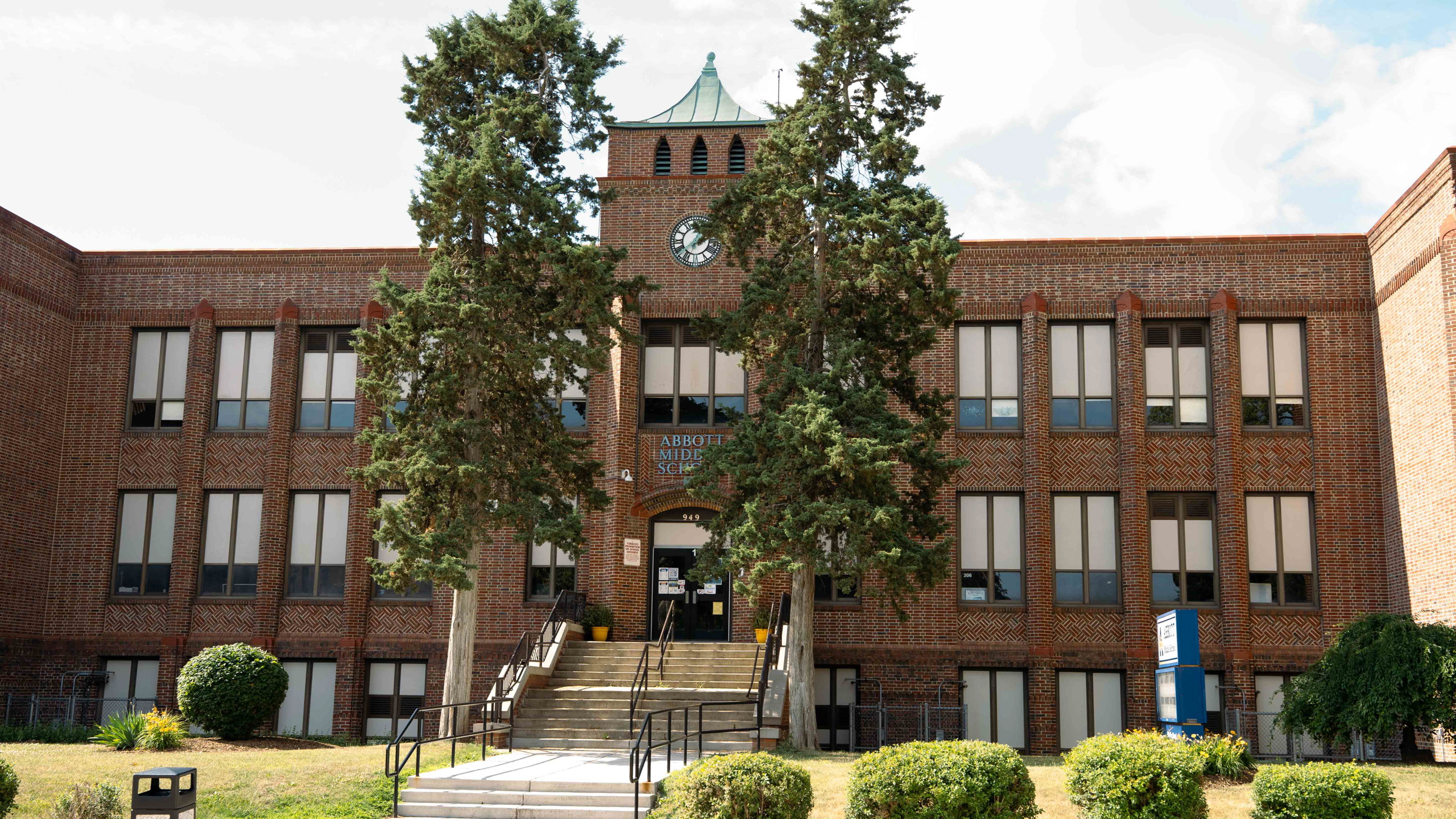A large brick building with multiple windows and a clock tower. It has trees and shrubs in the foreground.