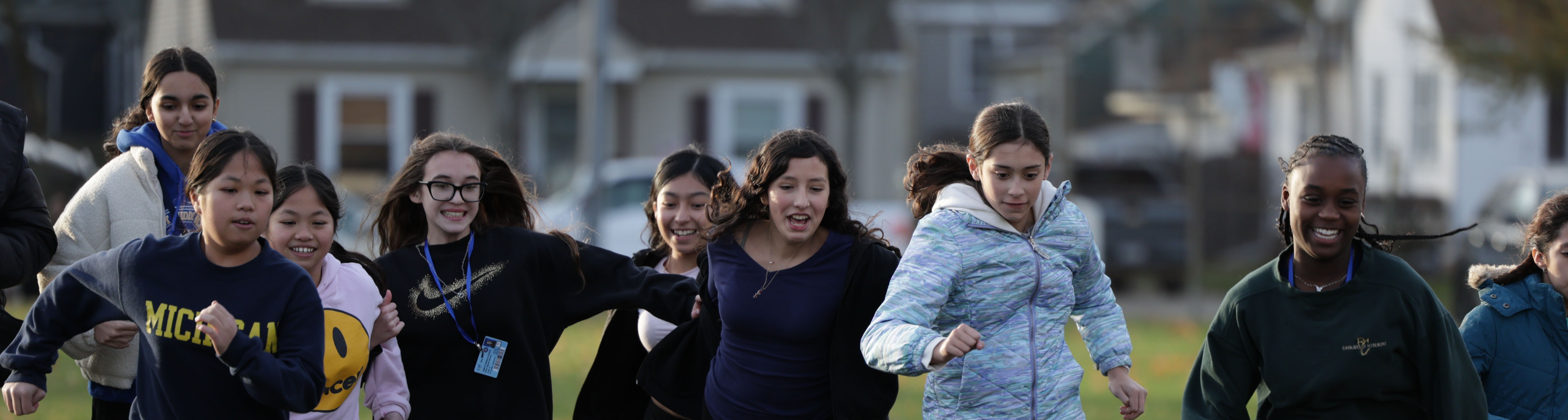 A group of people dressed in athletic clothing run on a grassy field, with houses visible in the background.